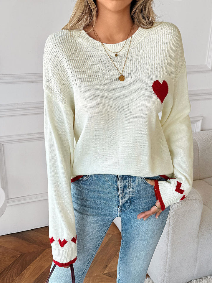 Person wearing a cream sweater with a red heart design and blue jeans indoors.