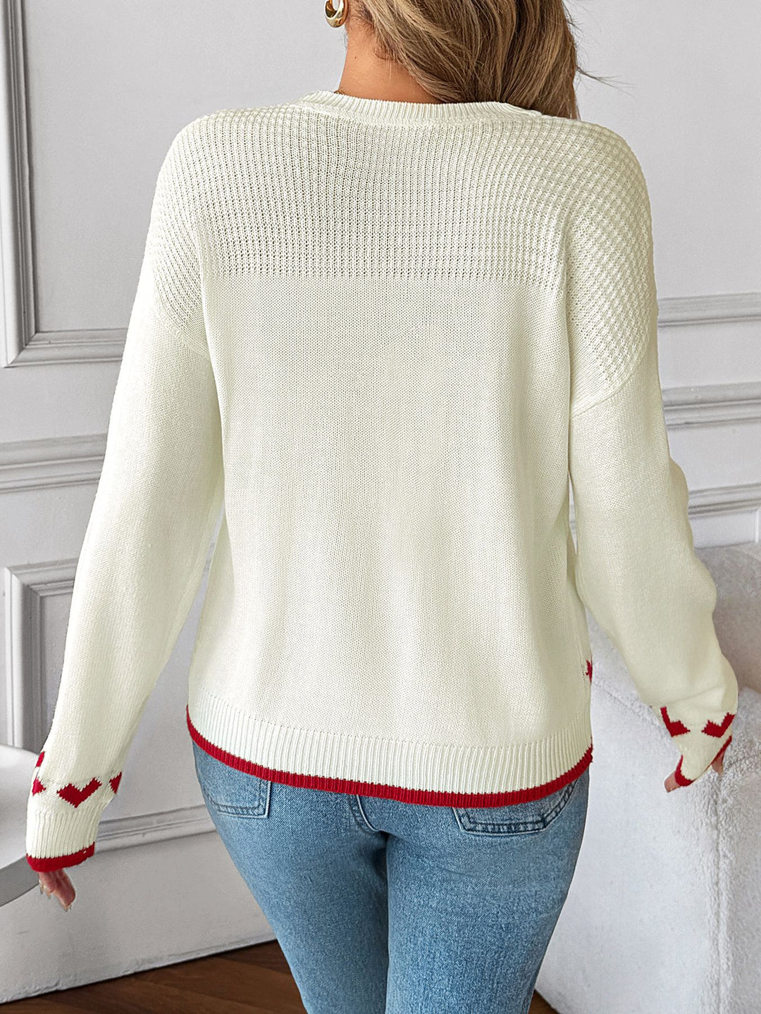 Person wearing a cream sweater with red accents and blue jeans against a gray wall.