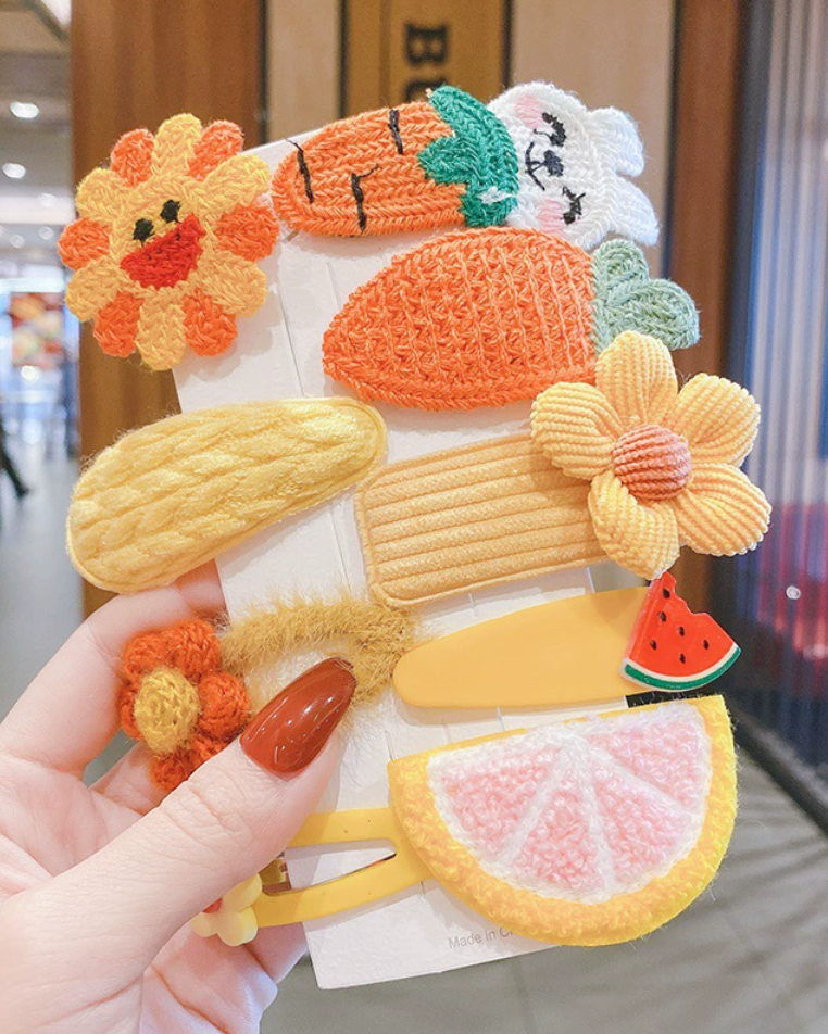 Colorful fruit-themed hair clips held by a hand in an indoor setting.