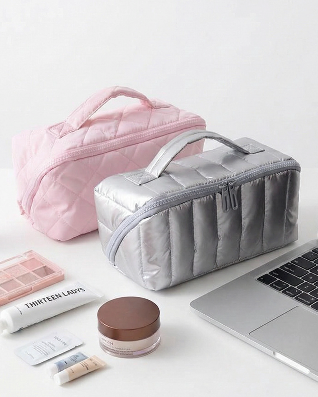 Gray toiletry bag next to a pink one on a white surface with a laptop and cosmetic items.