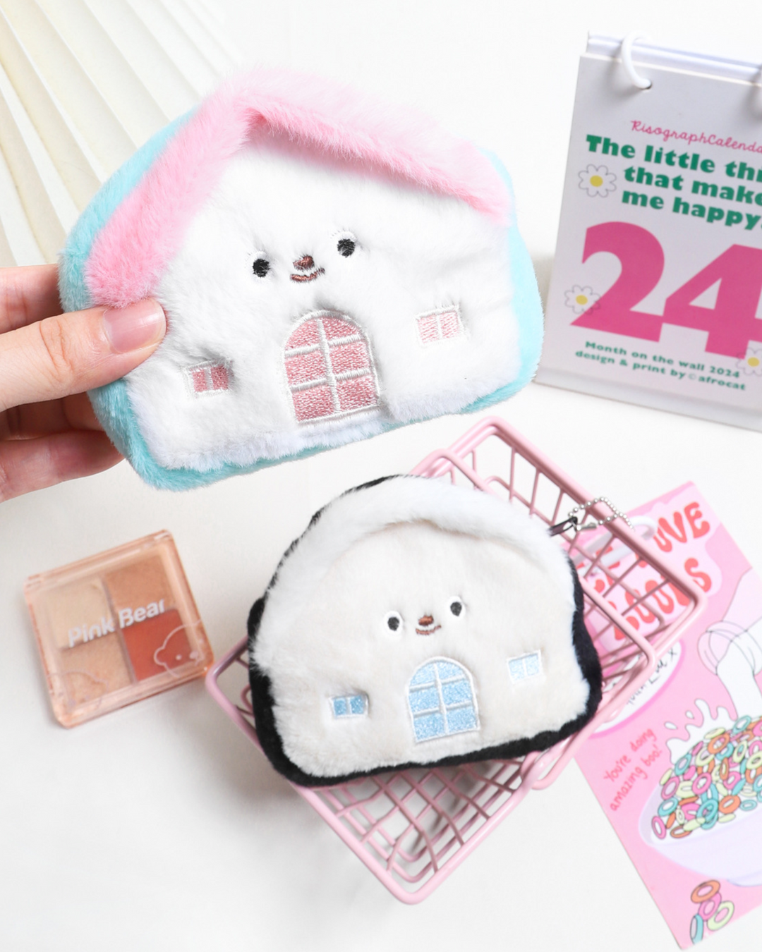 Small plush toy with a house design held by a hand, surrounded by stationery items on a white surface.